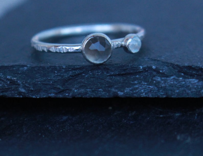 May include: A silver ring with a textured band features two gemstones: a larger, round, smoky quartz and a smaller, light blue stone. The ring is set against a dark, textured surface.