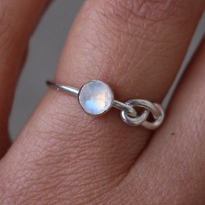 May include: A delicate silver ring featuring a small, round, iridescent moonstone. The ring's band is thin, with a knot design on the side. The moonstone has a subtle play of colors, reflecting light. A minimalist piece of jewelry.