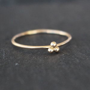 Könnte beinhalten: Ein zarter Goldring mit einer Ansammlung von drei kleinen, runden Perlen. Das Ringband ist dünn und glatt, die Perlen befinden sich oben. Der Ring ist vor einem dunklen Hintergrund platziert.