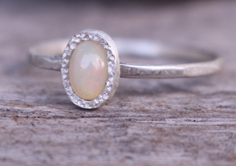 Puede incluir: Un anillo de plata con una piedra preciosa de &oacute;palo ovalada y blanquecina. El &oacute;palo est&aacute; engastado en un bisel texturizado. La banda del anillo es fina y tiene una textura martillada. El anillo se muestra sobre una superficie de madera.