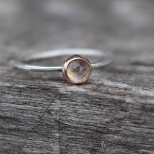 May include: A delicate ring with a silver band and a gold bezel setting. The ring features a round, light-colored gemstone. The band has a subtle dotted texture. The ring is set against a weathered wooden surface.