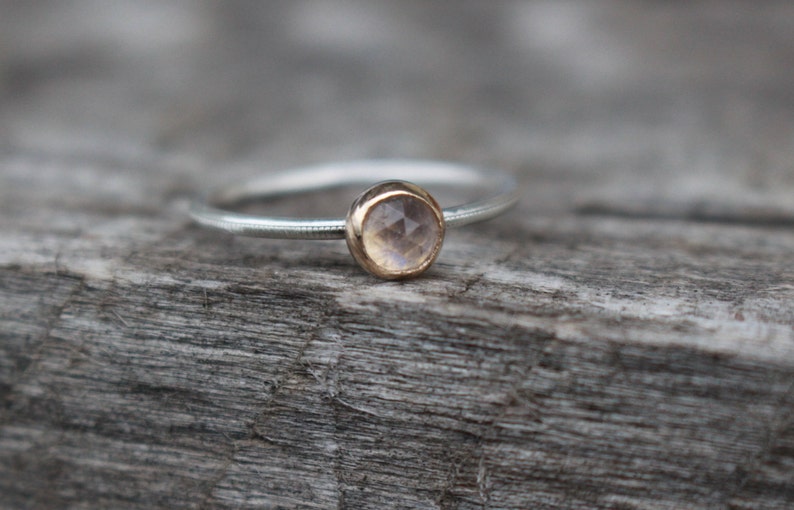 May include: A delicate ring with a silver band and a gold bezel setting. The ring features a round, light-colored gemstone. The band has a subtle dotted texture. The ring is set against a weathered wooden surface.