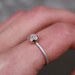 May include: A delicate ring featuring a small, round, faceted gemstone set in a gold bezel. The ring's band is silver with a textured, ridged design. The ring is shown on a finger, with a blurred background.