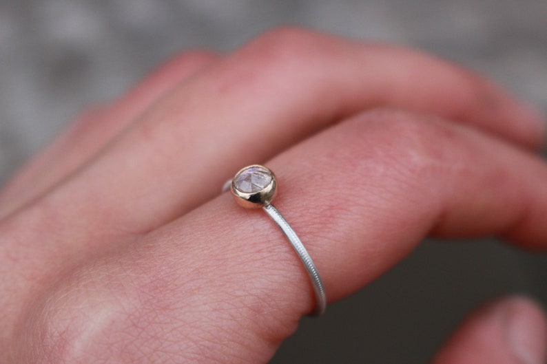 May include: A delicate ring featuring a small, round, faceted gemstone set in a gold bezel. The ring's band is silver with a textured, ridged design. The ring is shown on a finger, with a blurred background.