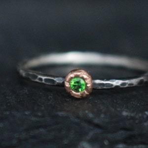 Puede incluir: Un anillo de plata con una banda texturizada martillada y un bisel de oro rosa. El bisel contiene una pequeña gema verde brillante y redonda. El anillo está sobre un fondo oscuro.