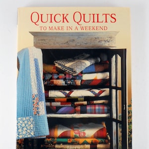 May include: A book titled "Quick Quilts to Make in a Weekend" with a cover featuring a display of colorful quilts. The quilts are stacked on shelves inside a dark-framed cabinet. A light blue quilt hangs on the left side of the cabinet.