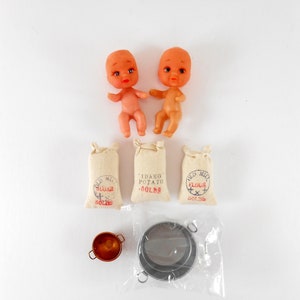 May include: A set of miniature dollhouse items including two plastic baby dolls, three fabric sacks labeled "Old Mill Sugar 50lbs", "Idaho Potato 50lbs", and "Old Mill Flour 50lbs", a copper pot, and a silver strainer.