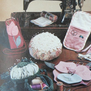 May include: A vintage sewing machine with a wooden base and a pink and white pincushion. There are also several other sewing accessories, including spools of thread, scissors, and a needle case.