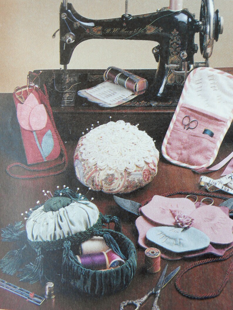 May include: A vintage sewing machine with a wooden base and a pink and white pincushion. There are also several other sewing accessories, including spools of thread, scissors, and a needle case.