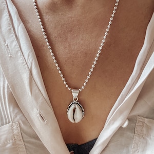 May include: Silver ball chain necklace with a cowrie shell pendant. The pendant features a white shell with a silver border and a detailed design. The necklace is displayed against a light-colored shirt.