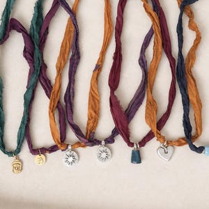 May include: A collection of seven necklaces featuring frayed silk ribbons in shades of teal, purple, mustard, and navy blue. Each necklace includes a unique metal charm, including a gold Buddha, an Om symbol, starbursts, a blue bead, and a heart.