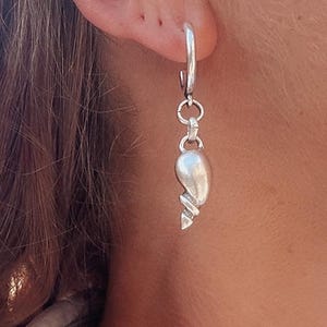 May include: A single silver earring with a teardrop-shaped pendant. The pendant has a twisted design at the bottom. The earring is attached to a hoop that is inserted into the ear.