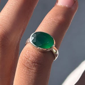 Peut inclure: Une bague en argent ornée d'une grosse pierre précieuse verte émeraude de taille ovale. La bague a un anneau en argent poli et la pierre est sertie dans un sertissage à lunette. La bague est portée sur un doigt.