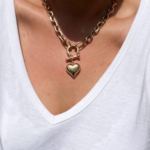 May include: Gold-coloured chain necklace with a heart pendant. The necklace has a chunky chain design and a toggle clasp. The heart pendant is smooth and polished, reflecting light. The necklace is worn against a white v-neck top.