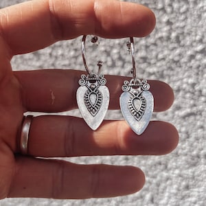 May include: A pair of silver-coloured hoop earrings. Each earring has a heart-shaped pendant with an ornate design. The pendants have a detailed, textured surface. The earrings are held up by a hand.