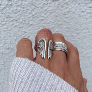 May include: A silver ring featuring a detailed wing design. The ring is worn on a finger with a second silver ring with a textured design.
