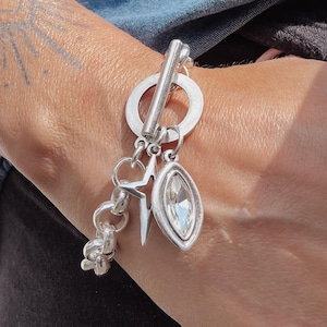 May include: Silver charm bracelet with a toggle clasp and various charms. Charms include a star, a crystal-like oval, and a knot design. The bracelet is worn on a wrist, with a denim shirt visible in the background.