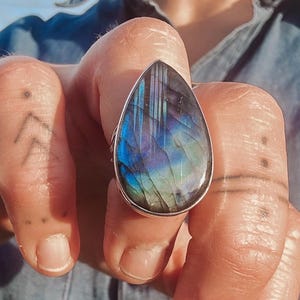 Può includere: Un anello d'argento con una grande pietra di labradorite a forma di goccia. La pietra presenta un gioco di colori vibranti, tra cui blu, verdi e viola. Un gioiello di grande effetto, che mette in risalto la bellezza naturale della gemma.