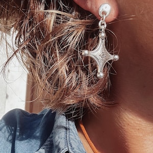 Può includere: Primo piano di un orecchino d'argento con un design a forma di croce. L'orecchino ha una piccola parte superiore rotonda e una forma a stella dettagliata a quattro punte con piccoli accenti a sfera. L'orecchino è indossato sull'orecchio di una persona con capelli corti e castani.