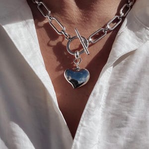 Könnte beinhalten: Eine silberfarbene Halskette mit einem Herzanhänger. Die Halskette hat eine klobige Kette und einen Knebelverschluss. Der Herzanhänger ist glatt und reflektierend. Der Schmuck wird vor einem weißen Hemd präsentiert.