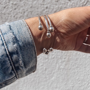 Könnte beinhalten: Silberne Armreifen mit runden Kugelakzenten. Die Armreifen sind auf dem Handgelenk einer Person gestapelt.