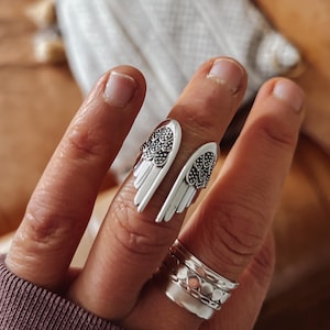 May include: A silver ring featuring a design of two angel wings. The wings are detailed with small circles and have a textured finish. The ring is worn on a finger with other silver rings.