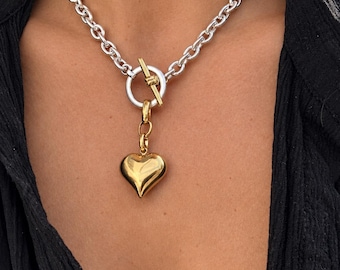 May include: A silver-toned chain necklace featuring a toggle clasp closure with a gold-toned bar. A polished, smooth gold-toned heart-shaped pendant hangs from the center of the toggle ring, creating a mixed-metal look with a classic link chain design.