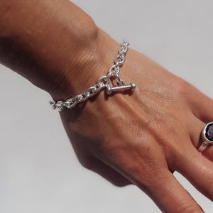 May include: A silver chain bracelet with a T-bar clasp is displayed on a person's wrist. The bracelet features a chunky link design. A silver ring with a dark purple stone is also visible.