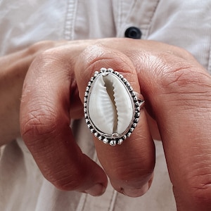 May include: A silver ring with a white and tan seashell centre. The ring has a beaded edge and is worn on a finger. The background is a light-coloured shirt.