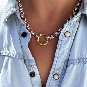 May include: A silver chain necklace with a gold ring pendant. The necklace is worn by a person wearing a light blue denim shirt.