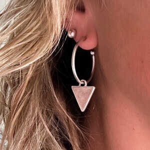 May include: Silver hoop earrings with a small triangle pendant. The earrings are worn on a person's ear, with the focus on the jewelry. The earrings are a simple, minimalist design.