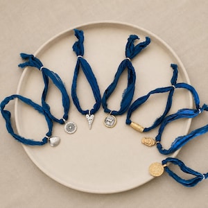 May include: A collection of seven bracelets made from dark blue frayed silk ribbons, each featuring a unique metal charm. The charms include a silver sun, a silver heart, a silver coin with a horse, a gold textured tube, and gold coin-shaped pendants.