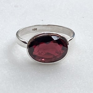 May include: A silver ring featuring a large, oval-cut, deep red gemstone. The ring band is smooth and polished, and the gemstone is set in a bezel setting. The ring is marked with "925".