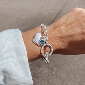 Puede incluir: Un brazalete de plata con un dije en forma de corazón y una piedra preciosa azul. El brazalete tiene un cierre de palanca.