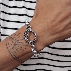 May include: A silver-toned bracelet featuring a prominent toggle clasp with a twisted rope-like texture. The bracelet is composed of thick, interlocking circular links that create a substantial chain design, showcasing a polished metallic finish.