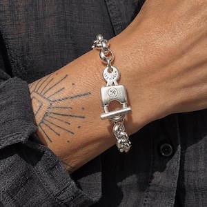 Op de afbeelding: Een zilveren kettingarmband met een knevelsluiting. De sluiting heeft de vorm van een hangslot met een cirkelvormig logo met de letters JH. De ronde schakel boven het slot is gegraveerd met de tekst MOGIUEC en 233.