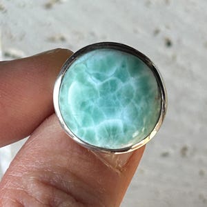 May include: A round ring featuring a polished, cabochon-cut larimar gemstone with a distinctive light blue and white marbled pattern, set within a smooth, plain silver-toned metal bezel that encircles the entire circumference of the stone.