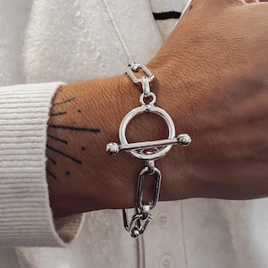Pode incluir: Uma pulseira prateada com fecho de alternância. A pulseira apresenta um design de elos de corrente com um fecho de alternância circular. A pulseira é usada em um pulso, com um suéter branco e uma camisa listrada ao fundo.