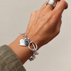 May include: A silver chain link bracelet featuring a toggle clasp, a smooth heart-shaped charm, and a small round blue stone charm, paired with a wide silver band ring with a textured, dotted pattern on the outer surface.