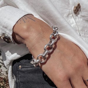 May include: A silver chain bracelet with large, rounded links. The bracelet is worn on a wrist, contrasting with a white long-sleeved shirt and dark denim jeans. The bracelet's design is bold and modern.