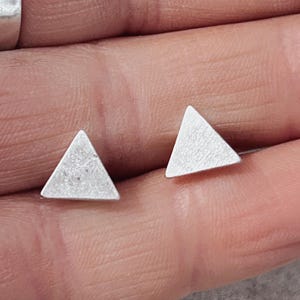May include: A pair of small, silver-colored stud earrings, each shaped as a simple equilateral triangle with a matte, brushed metal finish. The earrings feature a flat surface and clean, sharp edges, presenting a geometric design.