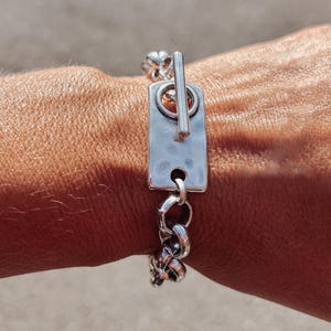 May include: Silver bracelet with a rectangular bar and toggle clasp. The bracelet has a chain-link design. The jewellery is displayed on a person's arm.