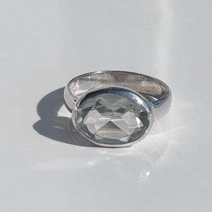 May include: A silver ring featuring a large, oval-cut, pale green gemstone. The ring band is smooth and polished, reflecting light. The gemstone is faceted, creating a sparkling effect. The ring is set against a white background.
