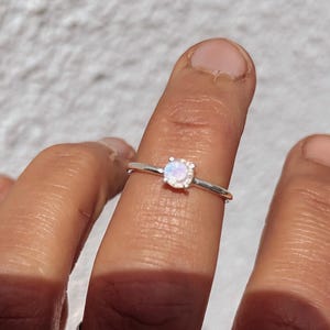 Op de afbeelding: Een zilveren ring met een ronde, iriserende opaal edelsteen. De ring is gezet op een dunne band en wordt op een vinger getoond. De opaal heeft een zachte, melkachtige uitstraling met kleurflitsen.