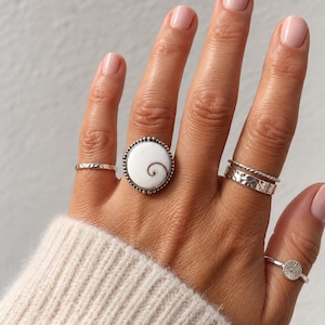 Könnte beinhalten: Ein Set aus fünf silberfarbenen Ringen an den Fingern. Der mittlere Ring zeigt ein großes, rundes, weißes Shiva-Auge-Muschel-Element mit braunem Spiralmuster in einer perlenartigen Silberfassung. Weitere Ringe sind ein dünner gehämmerter Ring, ein gedrehter Ring, ein breiter gehämmerter Ring und ein Ring mit Sonnenstrahlen-Motiv.