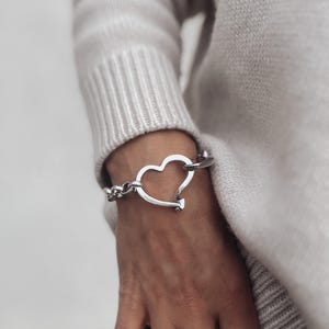 May include: A silver heart-shaped bracelet with a chain link design. The bracelet is worn on a person's wrist.