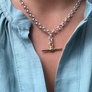Op de afbeelding: Een zilveren ketting met een goudkleurige staafhanger. De ketting is gemaakt van ronde, in elkaar grijpende schakels. De hanger is een horizontale staaf met een klein knoopdetail in het midden. De ketting wordt gedragen op een lichtblauw overhemd.