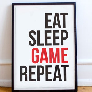 May include: A black framed poster with a white background and the text "Eat Sleep Game Repeat" in black and red lettering.
