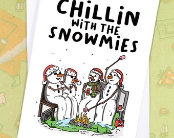 May include: A white greeting card with the text "Chillin' with the Snowmies" features a cartoon illustration of snowmen around a campfire. The snowmen wear Santa hats and scarves, roasting marshmallows. The card is on a white envelope.