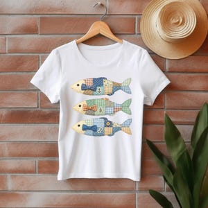 May include: White t-shirt featuring three colorful fish designs. Each fish has a patchwork pattern in blue, green, and yellow, with a bow tie and nautical-themed embellishments. The shirt is hanging on a wooden hanger.